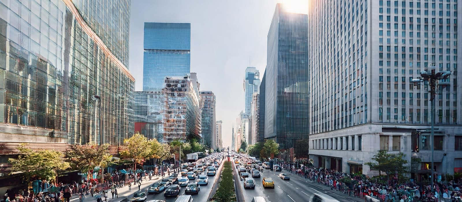 Wide, sunlit urban street lined with modern glass skyscrapers and heavy traffic, with pedestrians walking on the sidewalks.