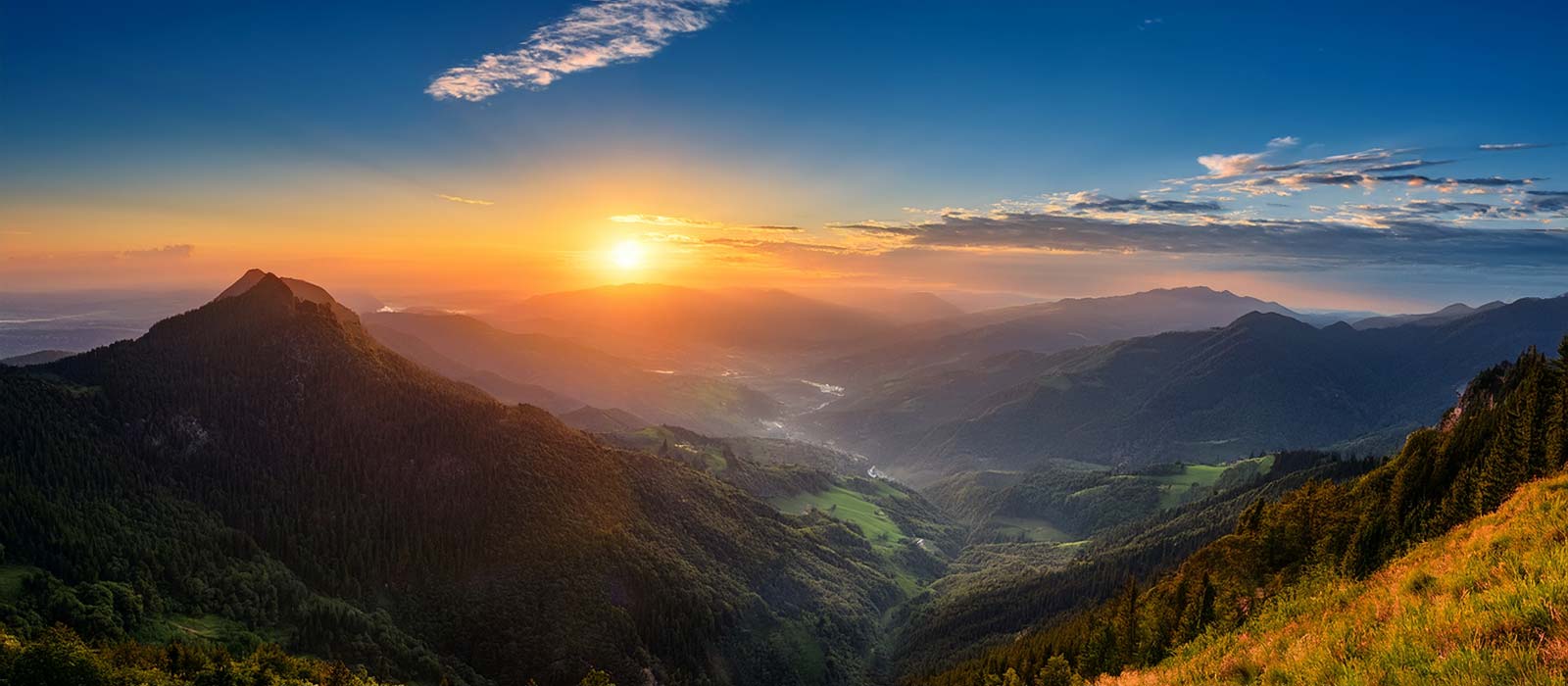 A vast mountain range at sunset, showing layers of forested hills and valleys under a brilliant orange and blue sky.
