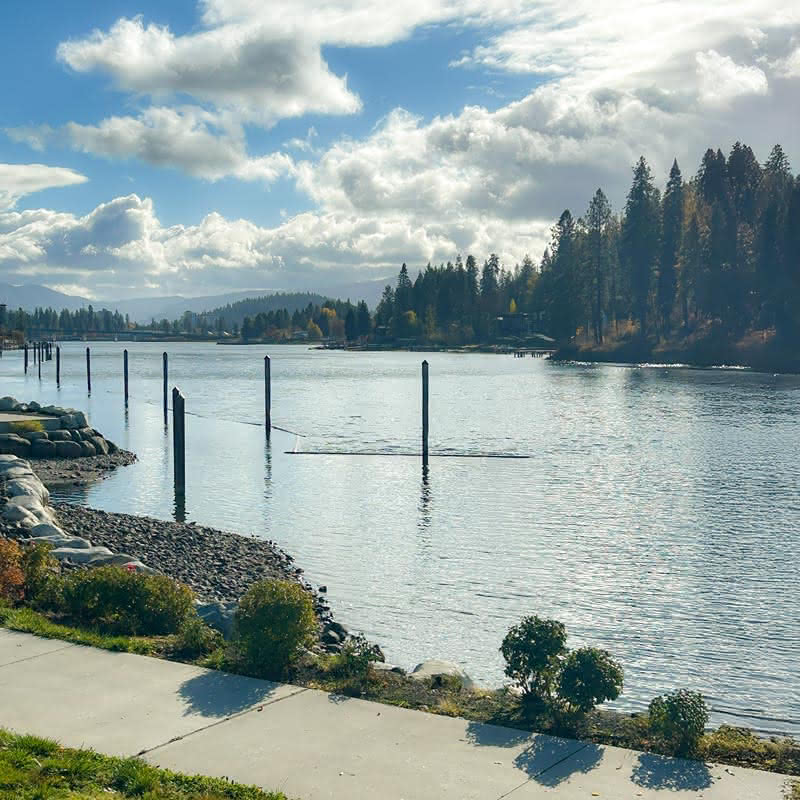 Wide view of a calm river with a shoreline path and dock posts in the water, framed by distant forested hills under a bright, cloudy sky.